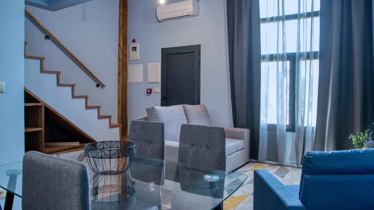 Sala com mesa de vidro, sofá azul e escada de madeira para o mezanino
